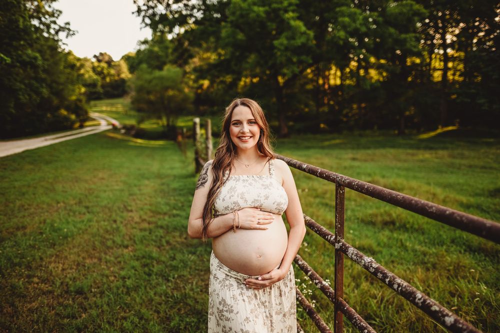 Expectant mother poses in lace dress by rustic fence during outdoor sunset photo session.