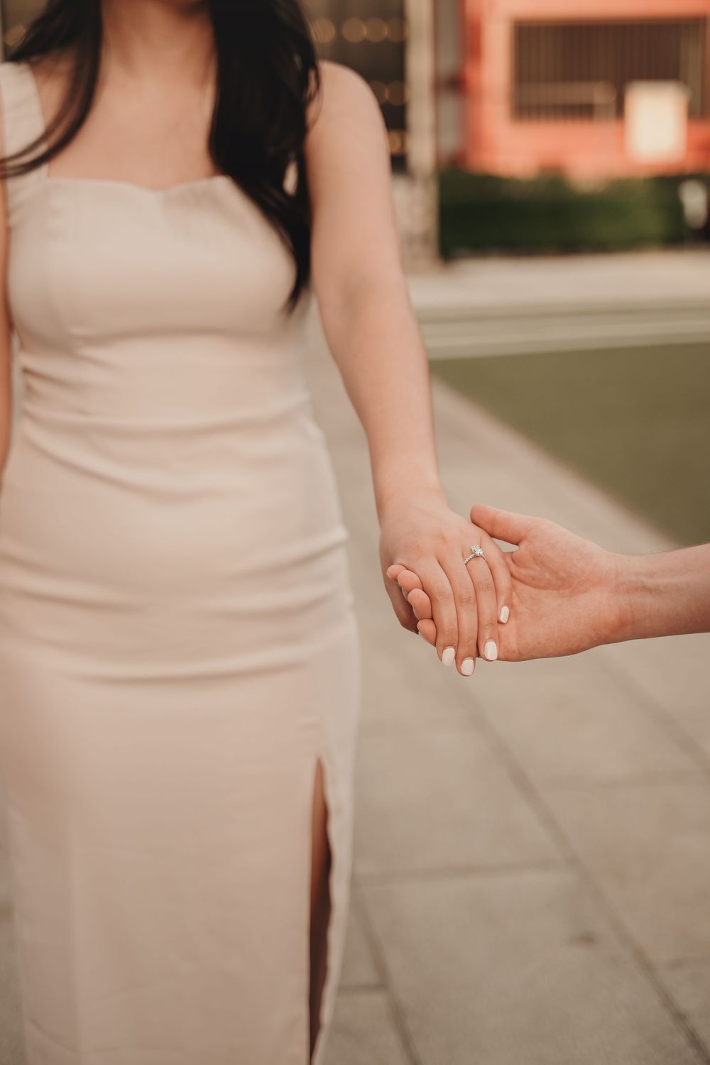 Couple holding hands outdoors, woman wearing elegant strapless beige dress with slit, engagement ring visible.