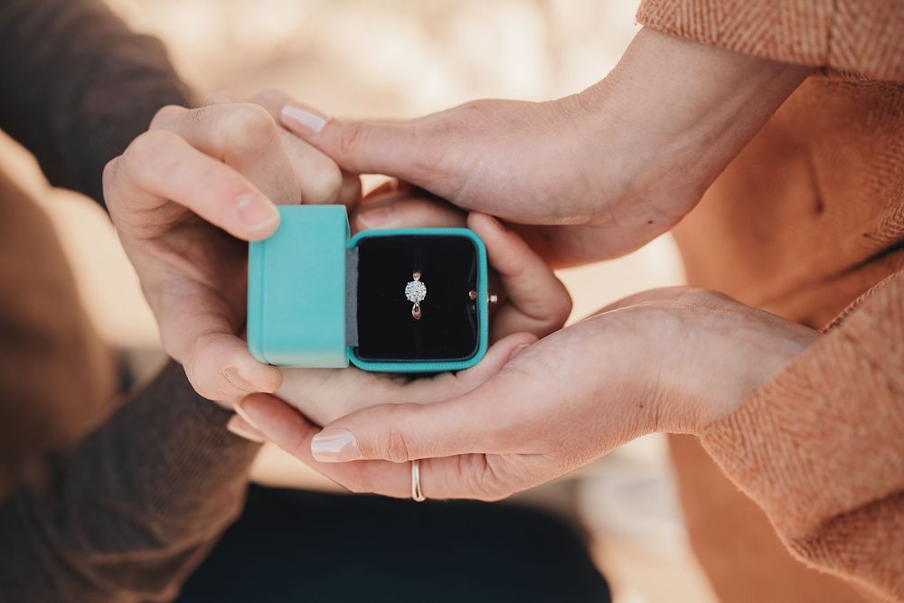 Two hands holding a teal ring box with a diamond engagement ring inside, symbolizing a romantic proposal.