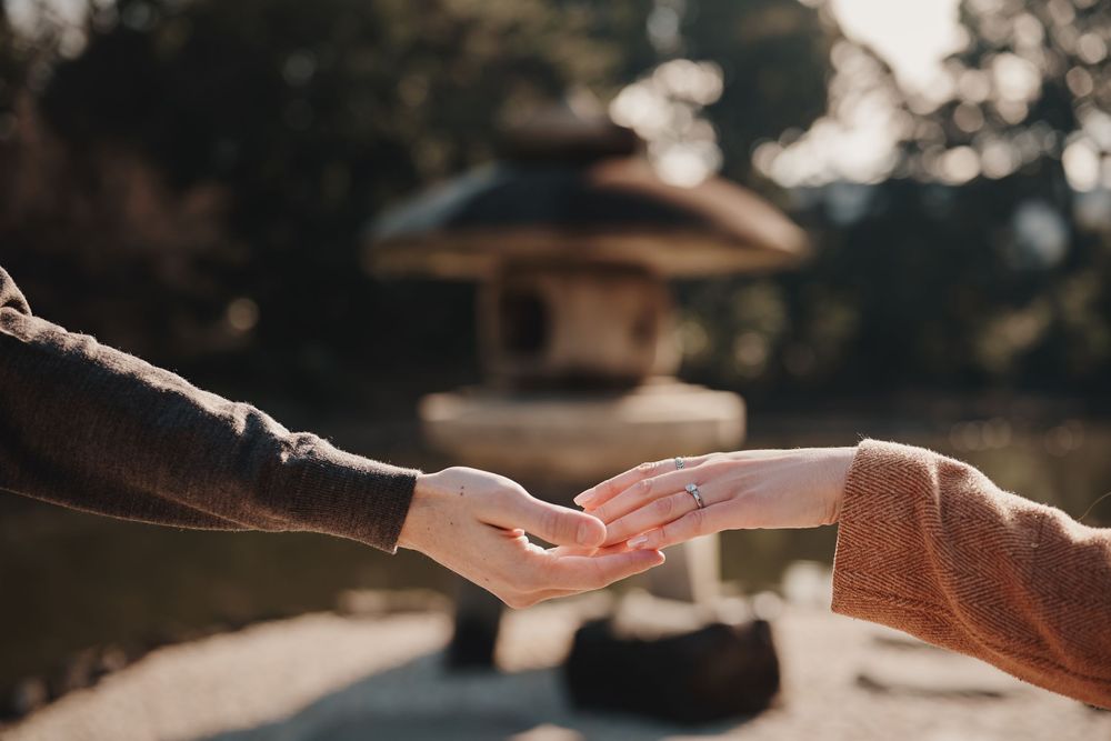 Two hands gently touching near a Japanese stone lantern in a serene garden, one wearing a diamond ring.