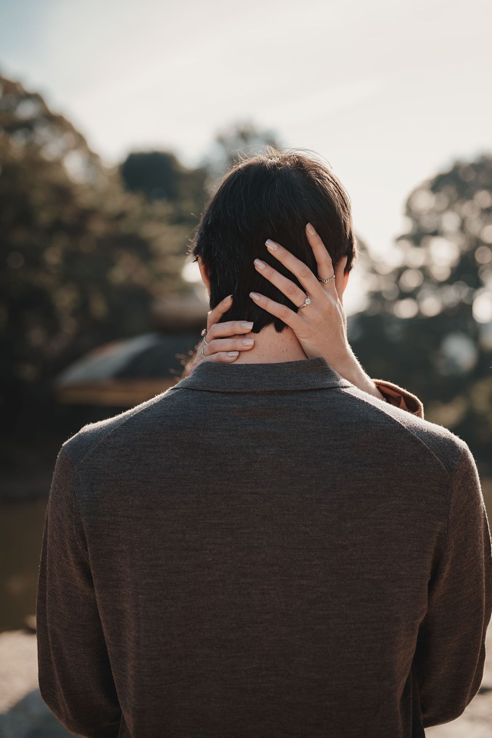 Back view of a person in a brown sweater with a hand gently placed on their neck outdoors.