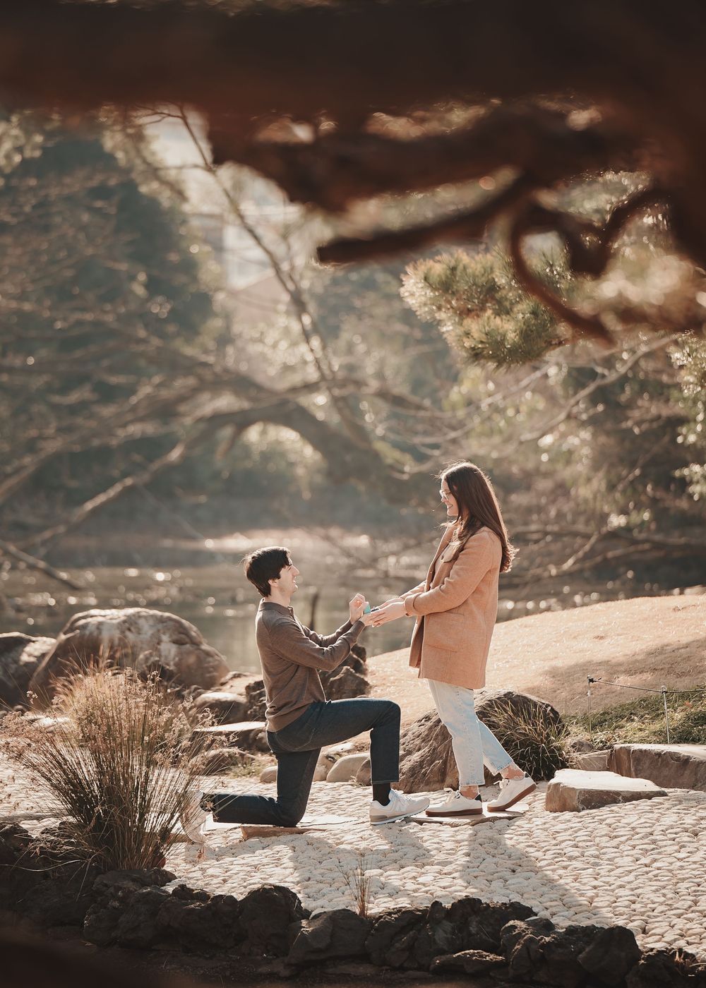 Man kneeling on one knee proposing to woman in pink coat outdoors in a scenic park setting.