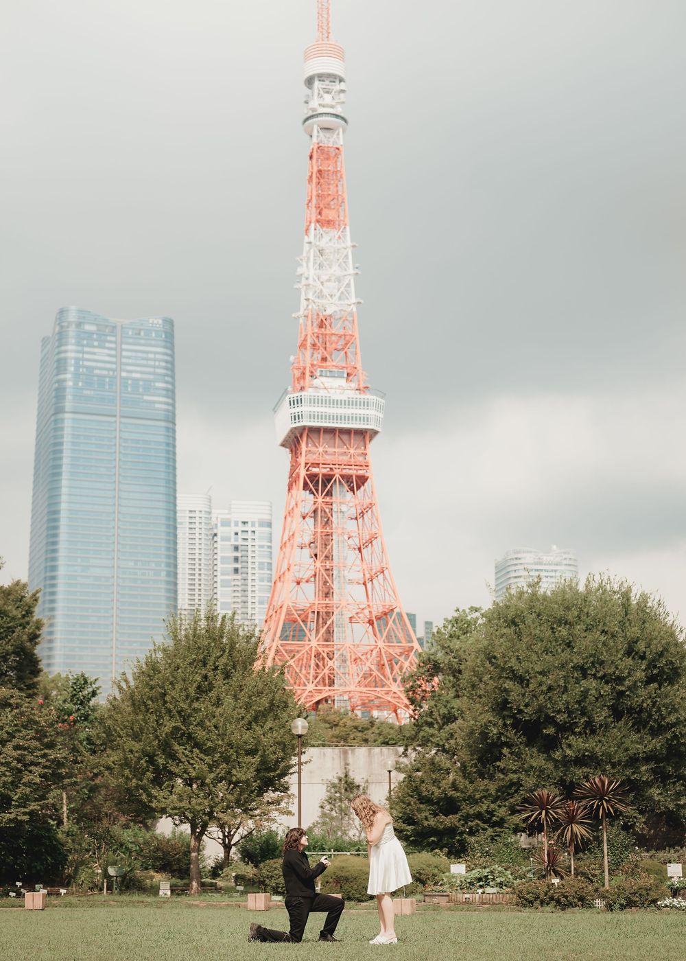 Couple's marriage proposal in front of Tokyo Tower with city skyline backdrop on a cloudy day.