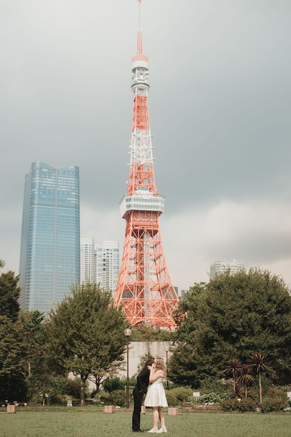 Couple embracing in front of Tokyo Tower with city skyline backdrop on a cloudy day.
