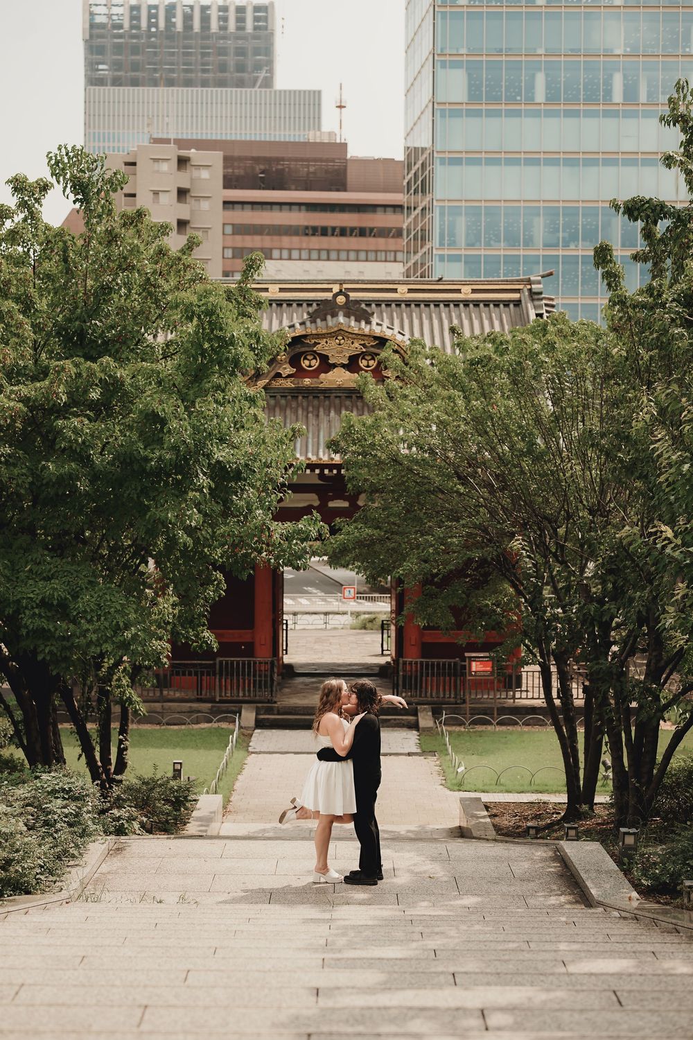 Couple sharing a romantic kiss in front of a traditional Japanese temple surrounded by lush trees and modern city buildings.