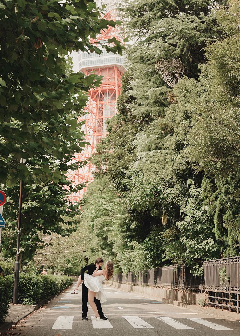 Couple sharing a romantic dip kiss on a crosswalk with Tokyo Tower and lush green trees in the background.
