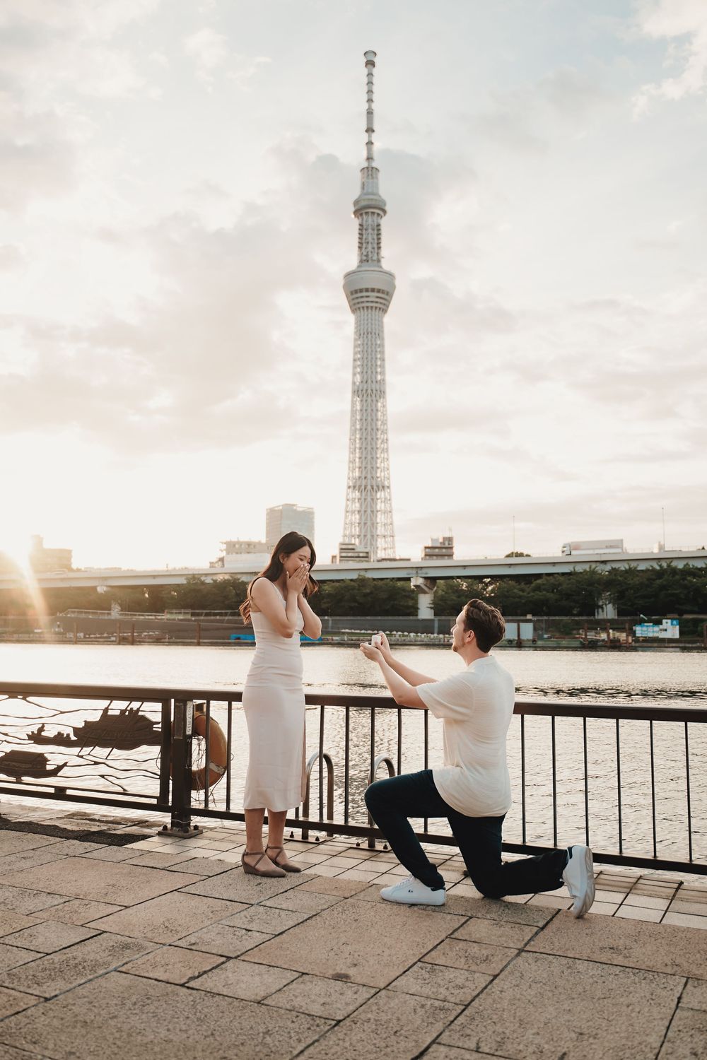 Man proposes to woman on one knee by a waterfront with Tokyo Skytree tower at sunset.