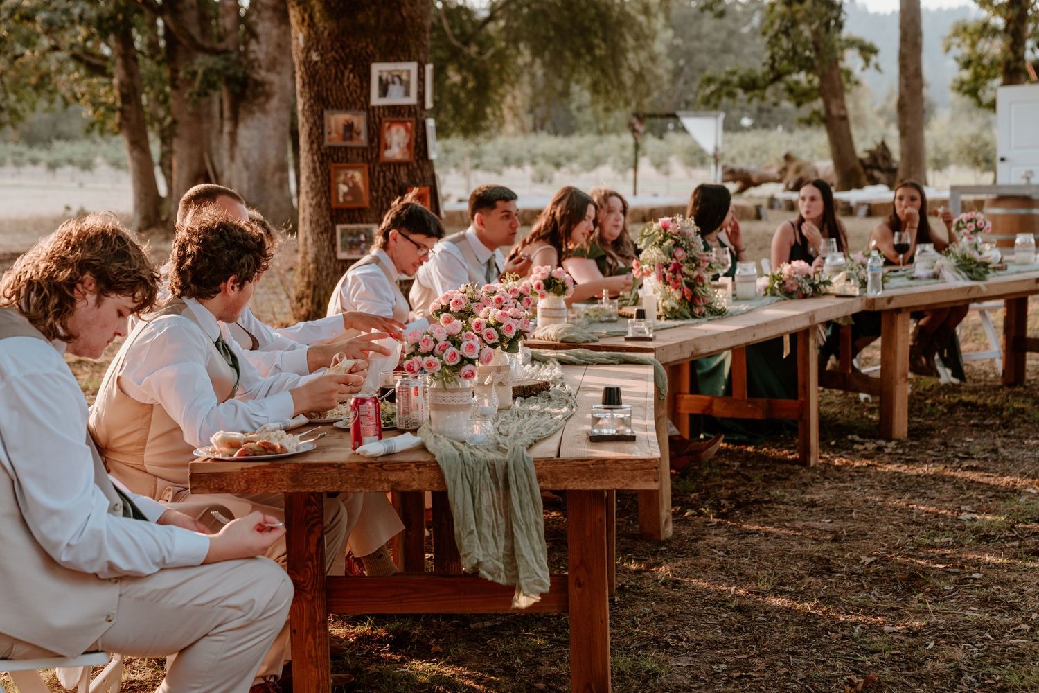 Outdoor wedding reception with guests seated at long rustic wooden tables decorated with pink floral arrangements.