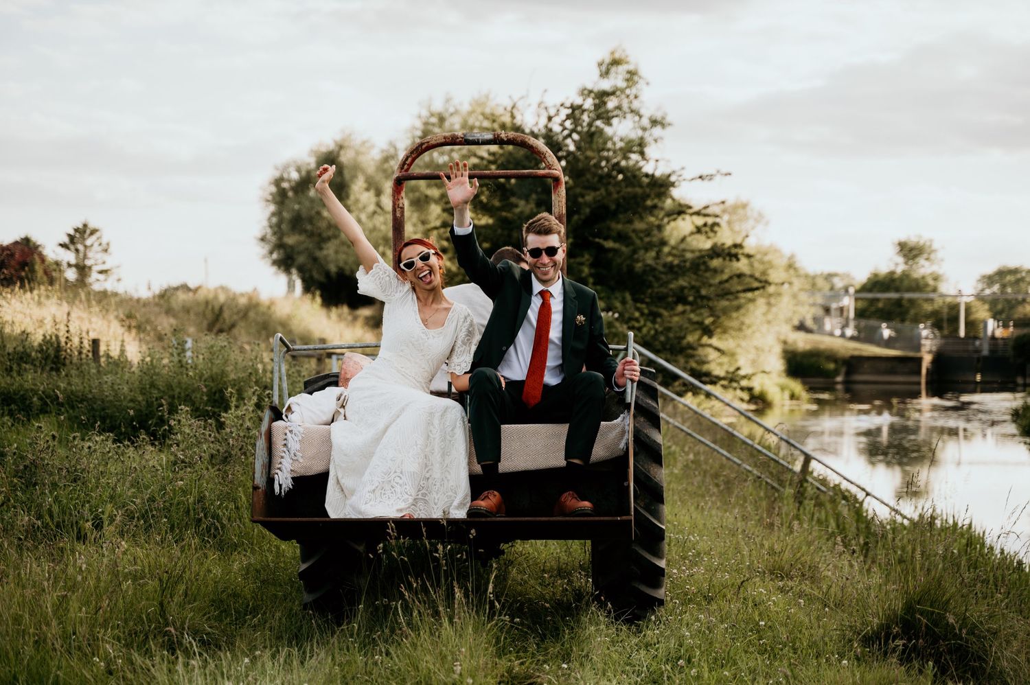 bride and groom sitting on the back of tractor next to river