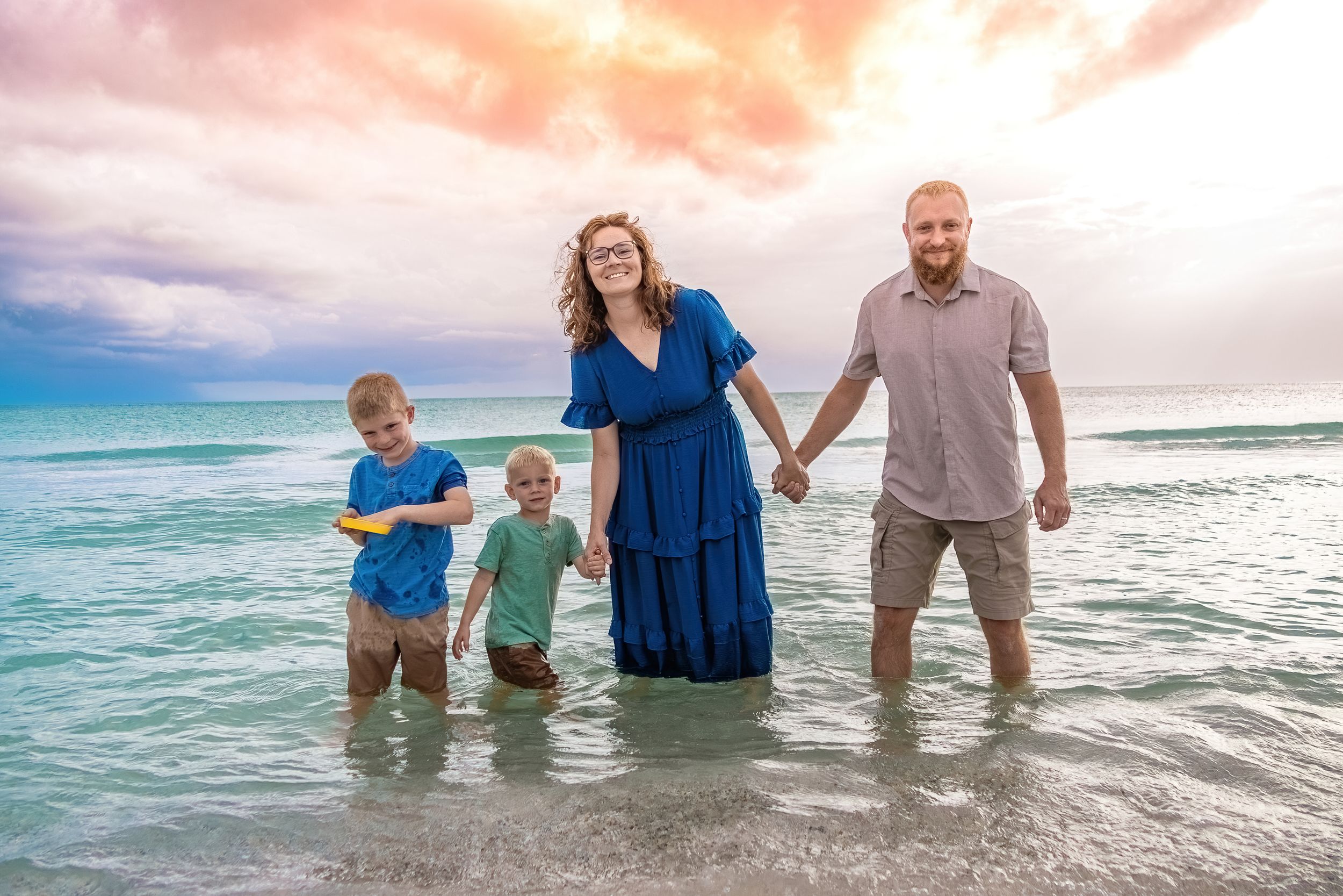 Family of four wading in ocean waves at sunset on a colorful beach, smiling and holding hands.