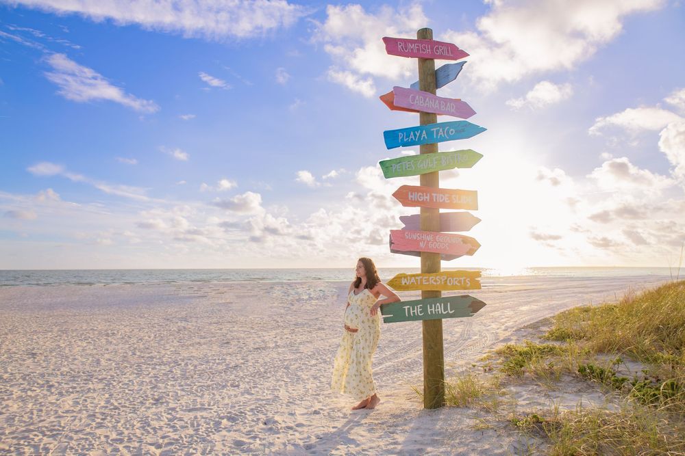 Woman in white dress standing by colorful directional signpost on pristine white sand beach with blue sky.