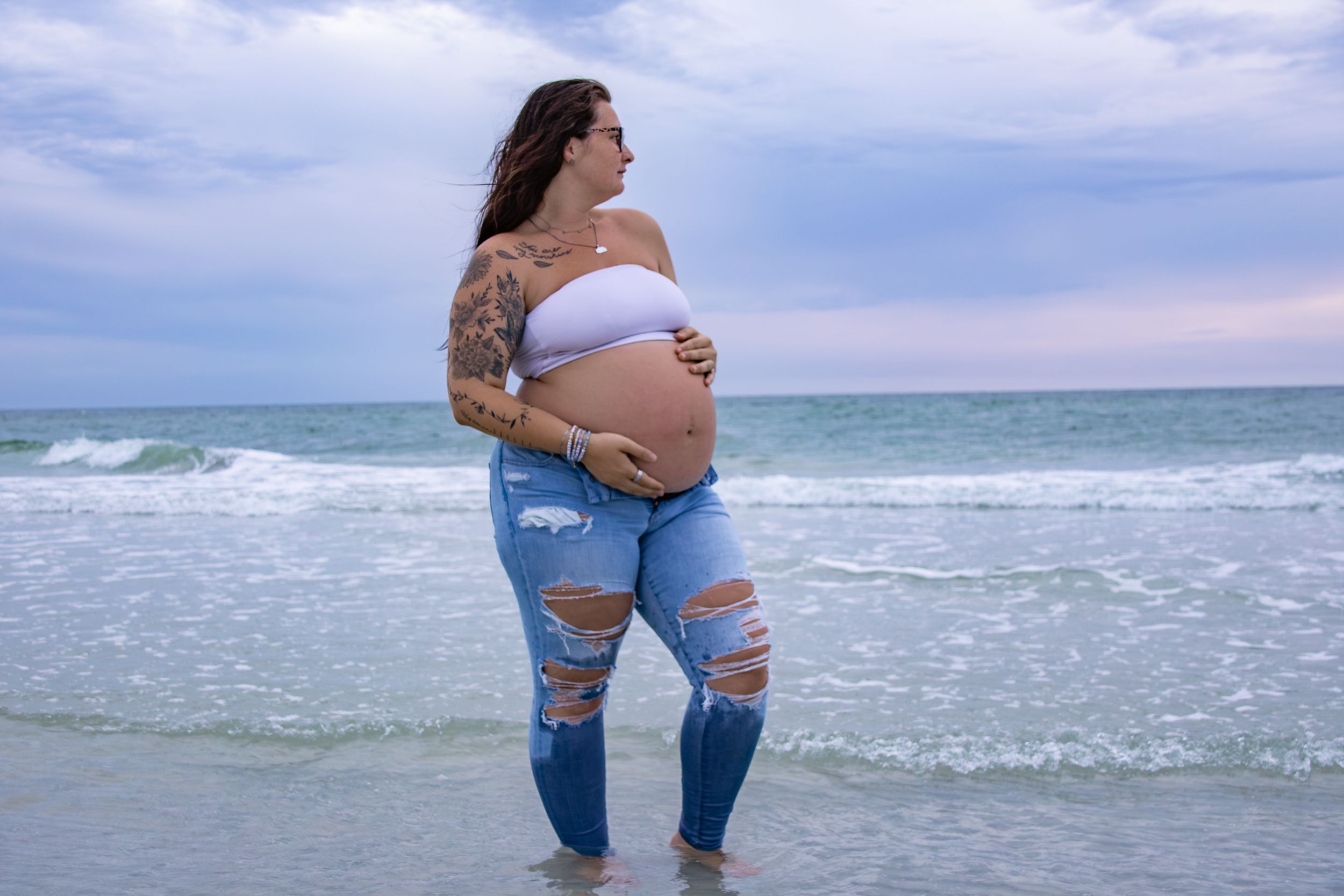 Pregnant woman with tattoos in ripped jeans and white crop top standing in ocean waves at beach.