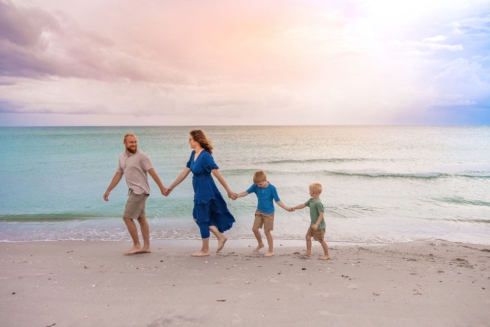 Family of four holding hands walking along a sandy beach at sunset with turquoise ocean waves.
