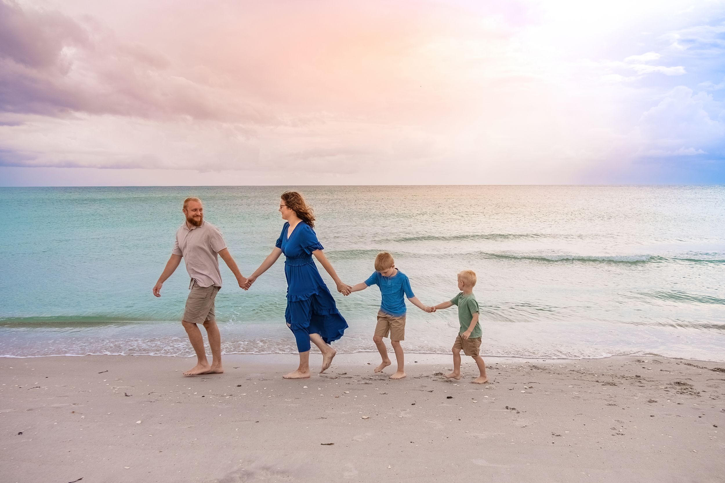 Family of four holding hands walking along a sandy beach at sunset with turquoise ocean waves.