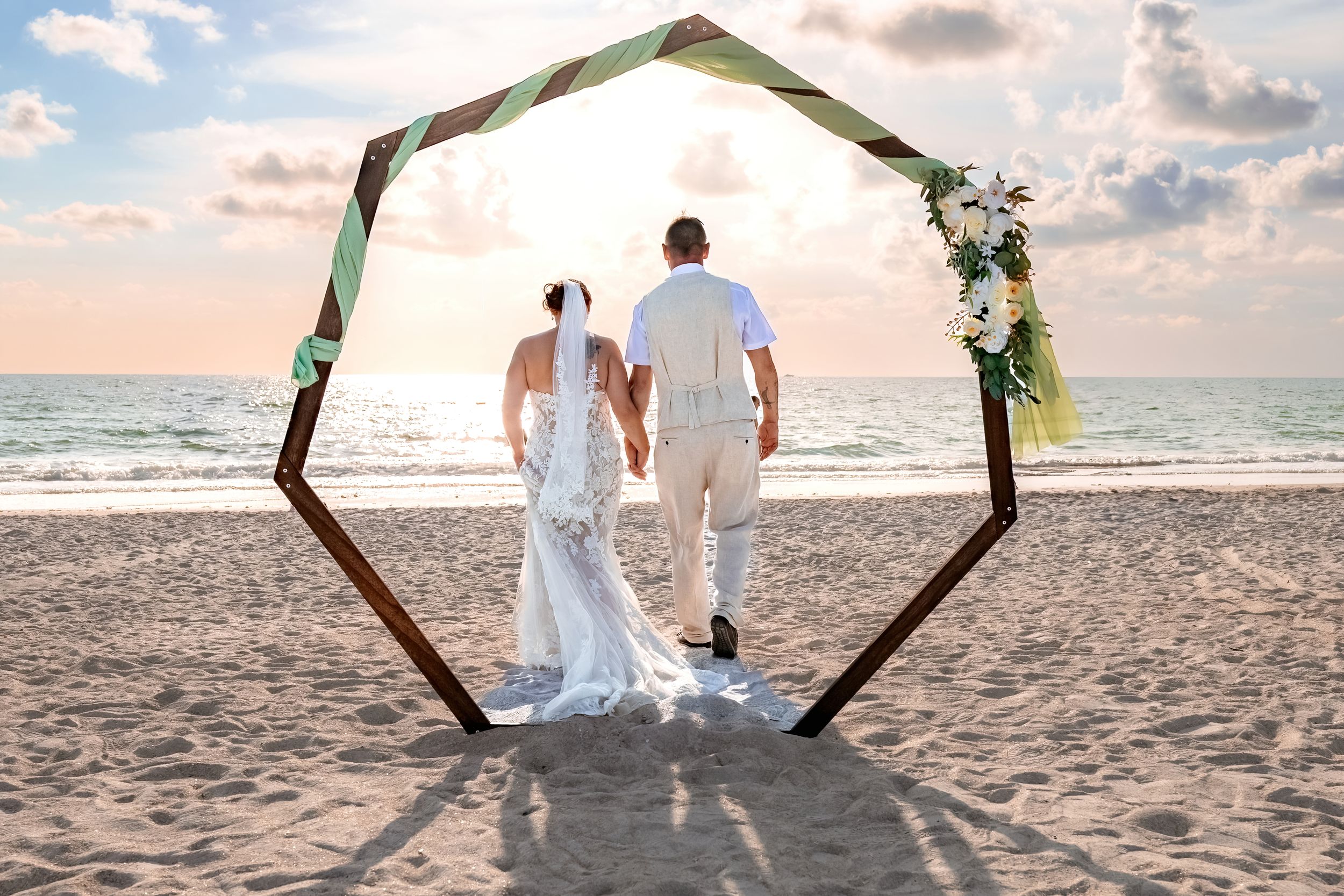 A couple stands beneath a hexagonal wooden arch decorated with greenery on a beach at sunset.