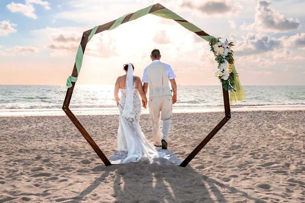 A couple stands beneath a hexagonal wooden arch decorated with greenery on a beach at sunset.