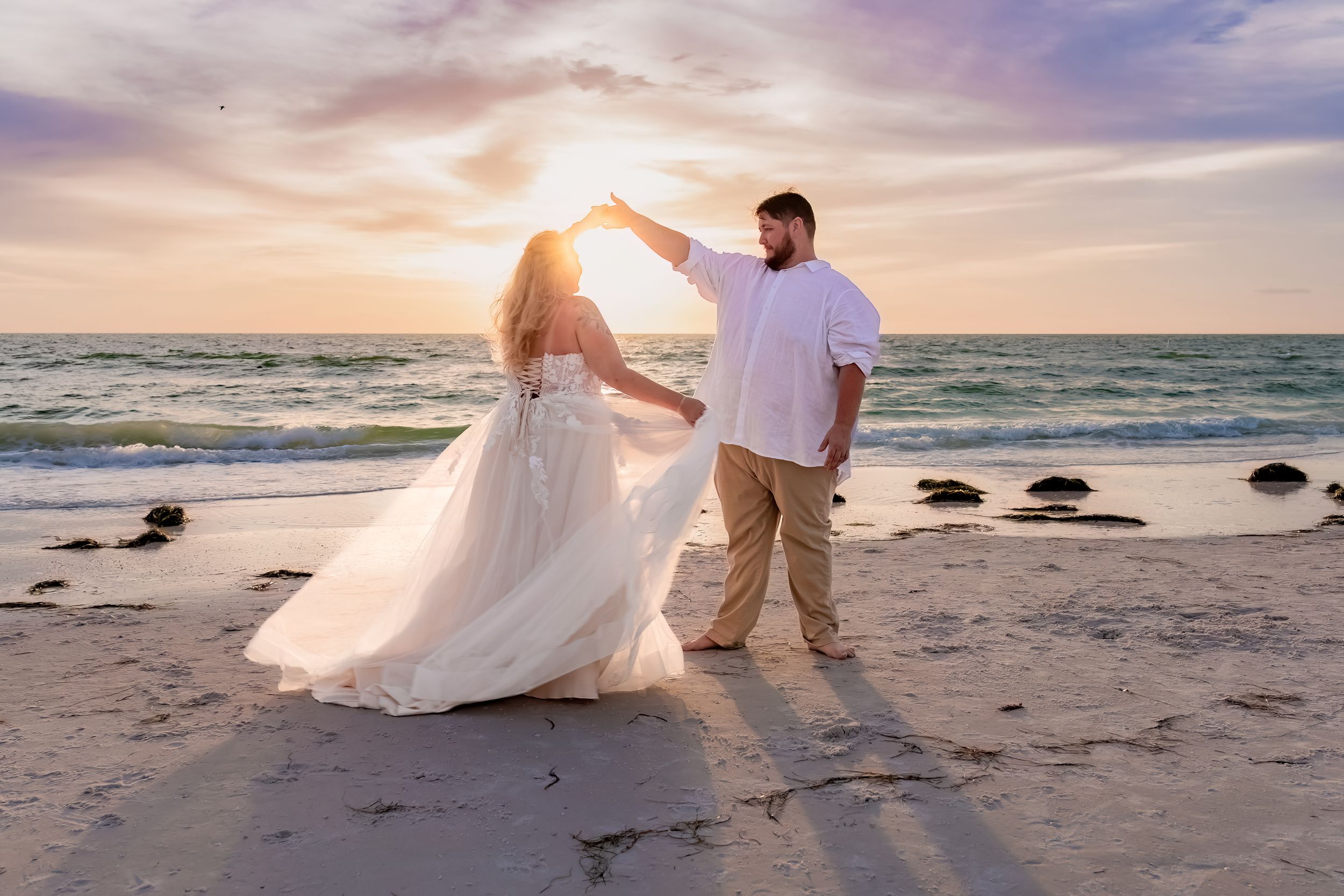 A couple dances together on a beach at sunset with waves crashing in the background and rocks along the shoreline.