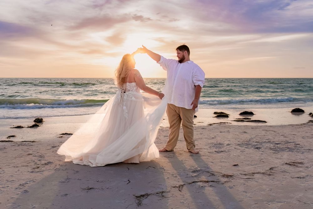 A couple dances together on a beach at sunset with waves crashing in the background and rocks along the shoreline.