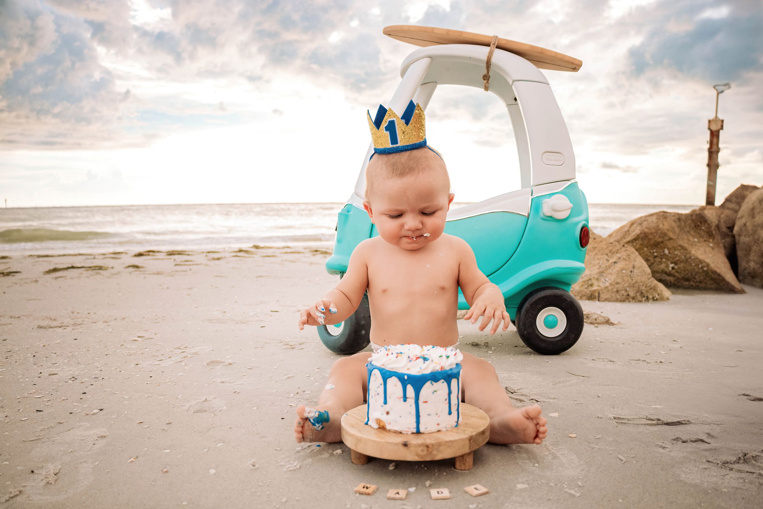 A baby in a blue cake smash photoshoot plays in frosting on a sandy beach at sunset.