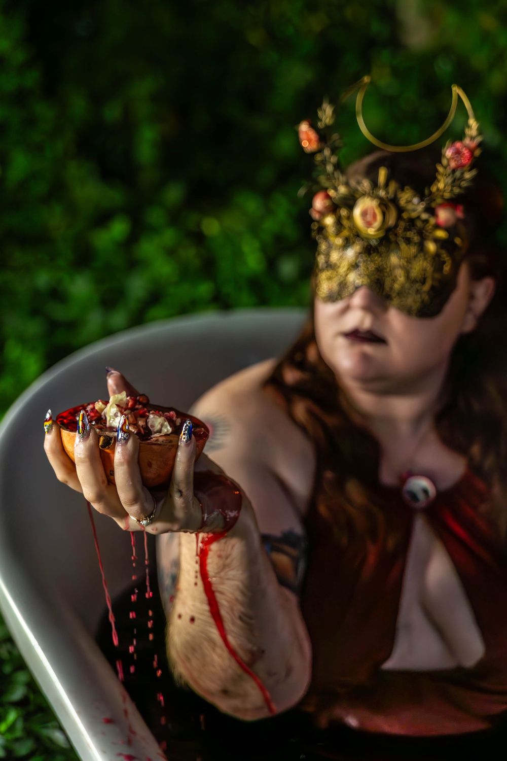 A woman dressed as Persephone baths in a red bath and squeezes a pomegranate.
