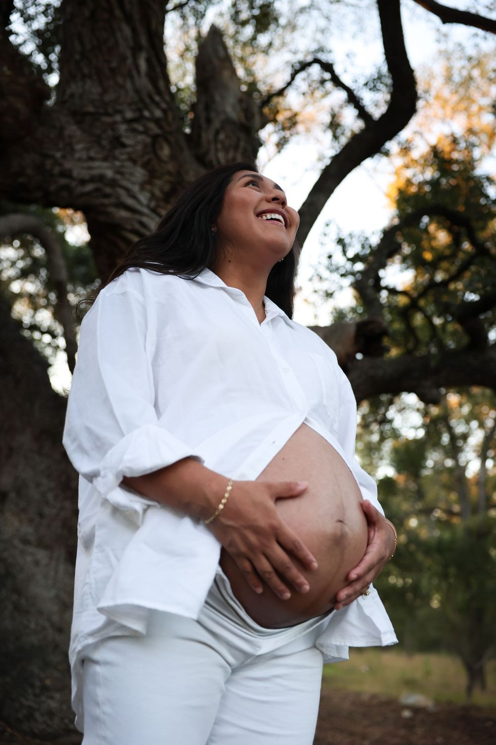 A pregnant person in a white dress cradles their belly while standing outdoors near trees at sunset.
