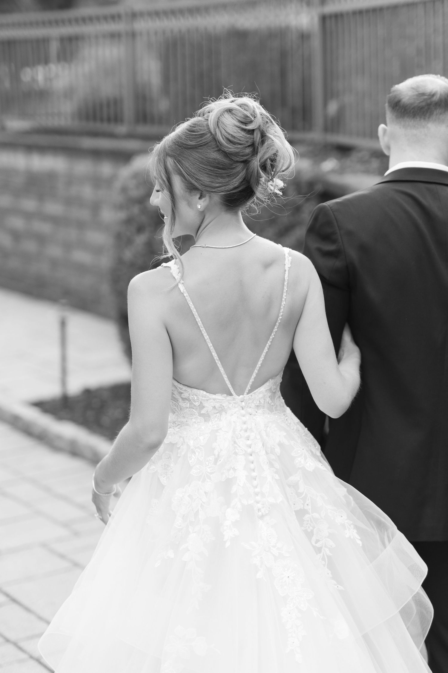 Black and white back view of bride in backless lace wedding gown with updo hairstyle walking with groom.