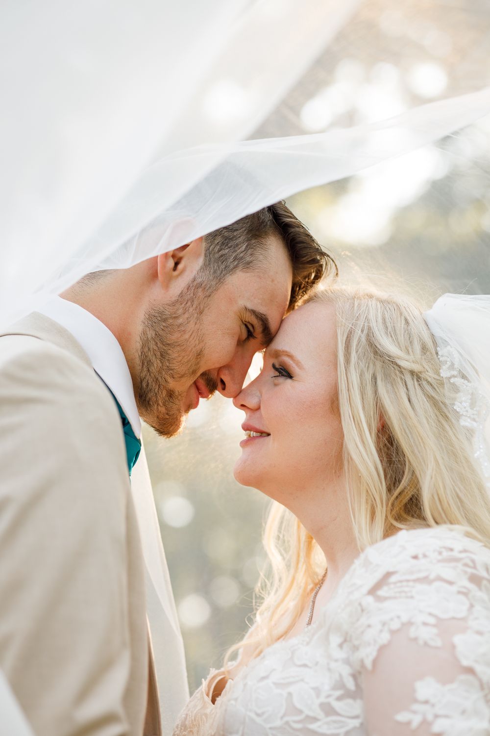 A tender romantic moment between newlyweds under a veil on their wedding day in warm sunlight.