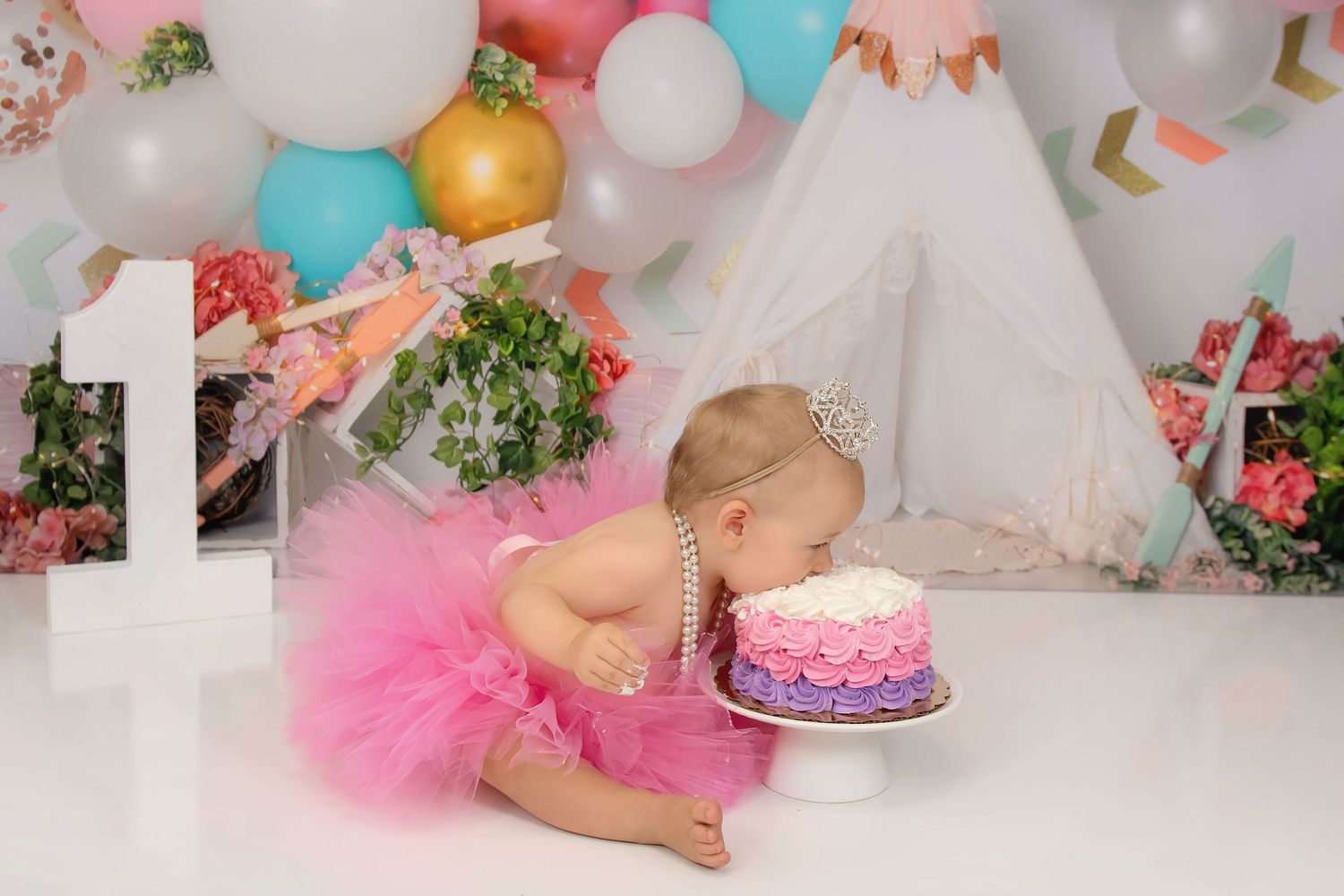 Baby in pink tutu leans over to take a bite of first birthday cake surrounded by balloons and decorations.