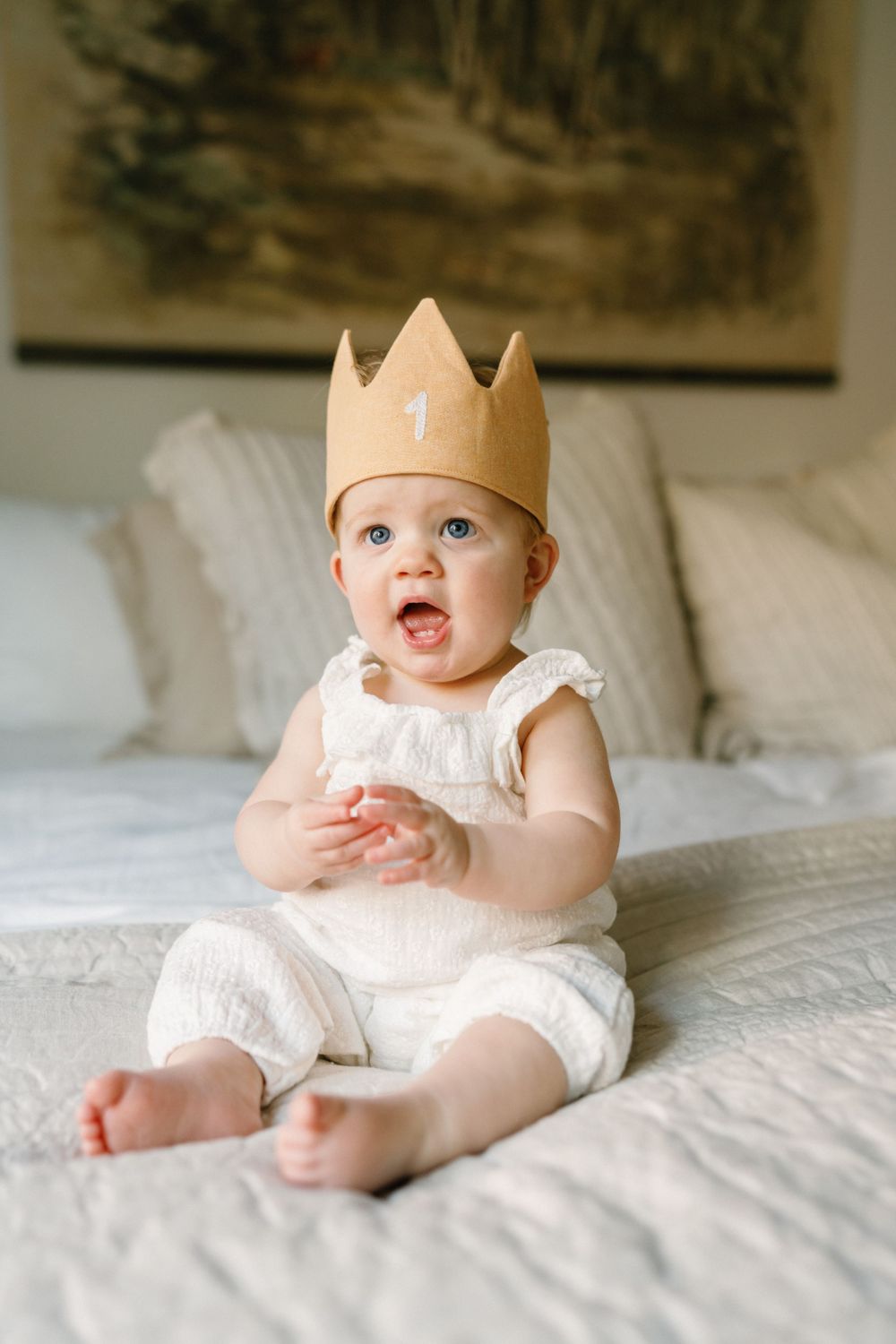 A young toddler in white clothing and a gold paper crown sits happily on a white bedspread.