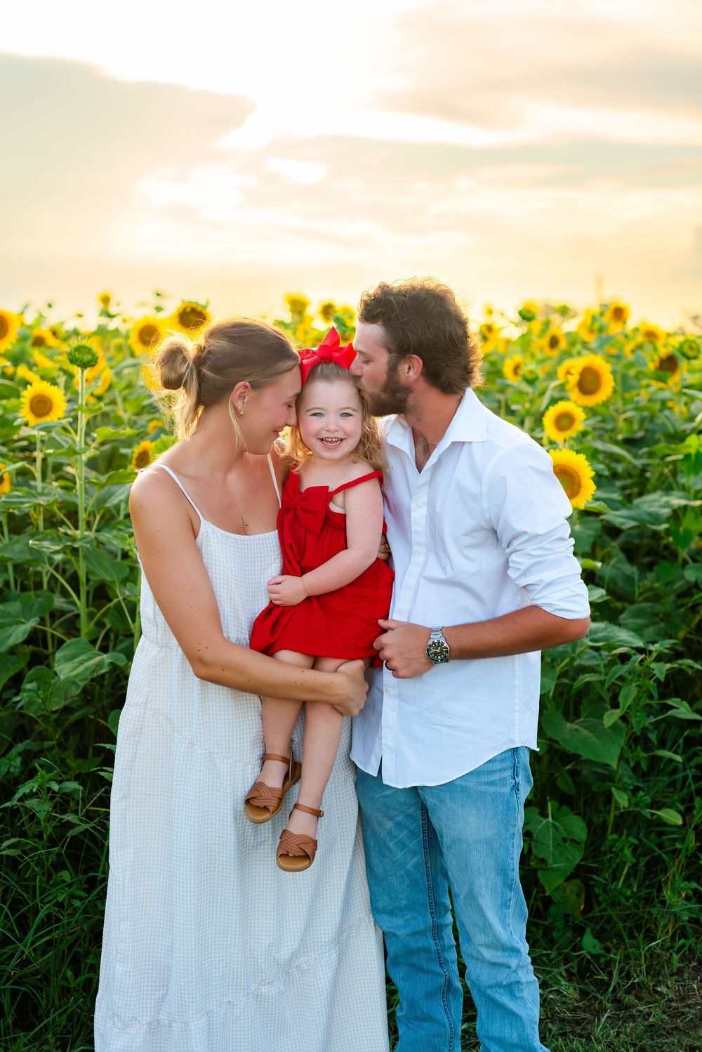 Three people embrace and share a joyful moment together in a scenic sunflower field during sunset.