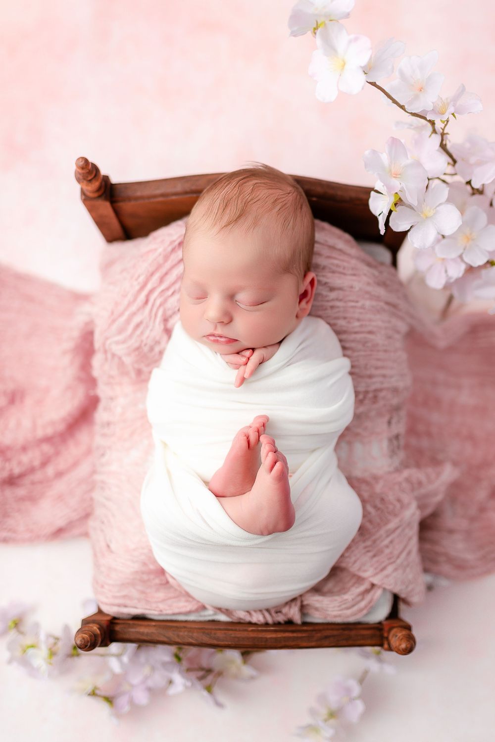 A sleeping newborn baby wrapped in white cloth lies on a soft pink blanket surrounded by delicate cherry blossoms.