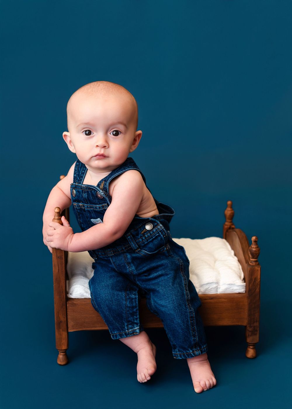 A baby wearing denim overalls poses on a small wooden bed against a dark blue background in a creative photo series.