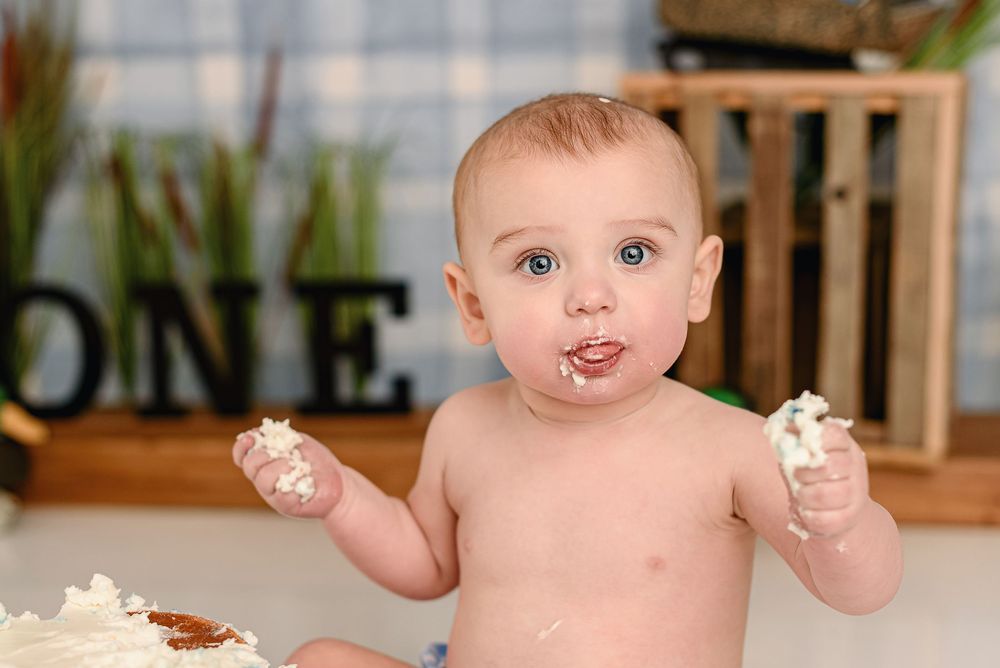 A baby with frosting on their hands reaches upward while sitting in a wooden chair by a window.