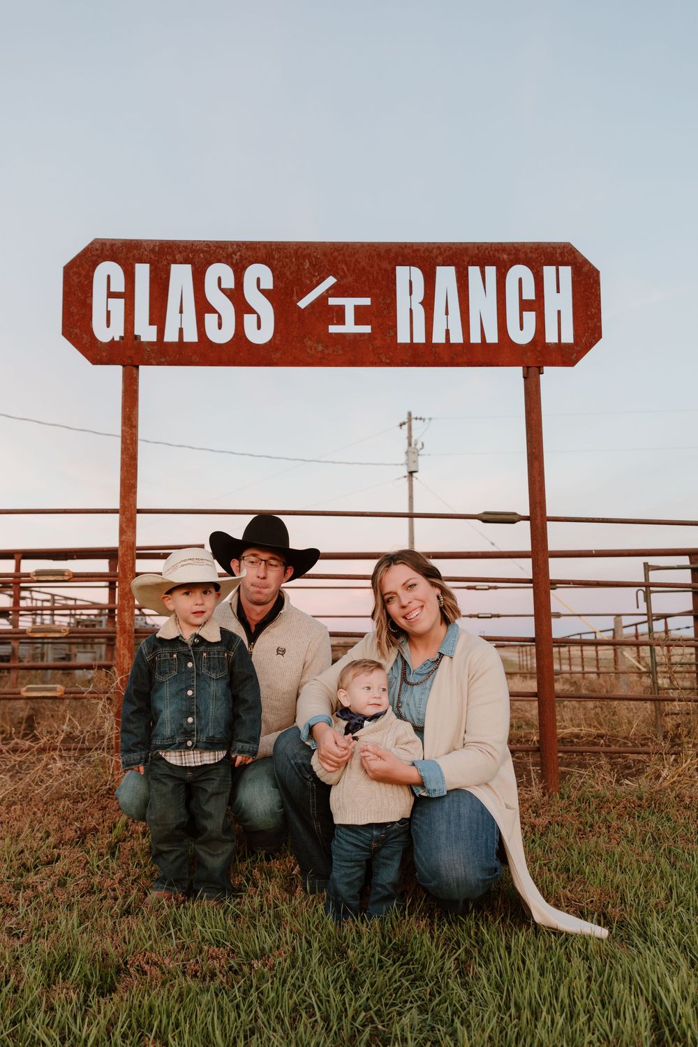 A family sits together in front of a rustic ranch sign and wooden fence at sunset.