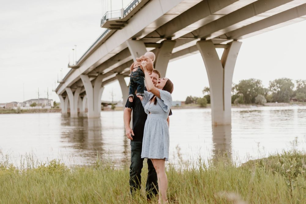 Romantic outdoor photoshoot with couple embracing under a bridge near water and tall grass at sunset.