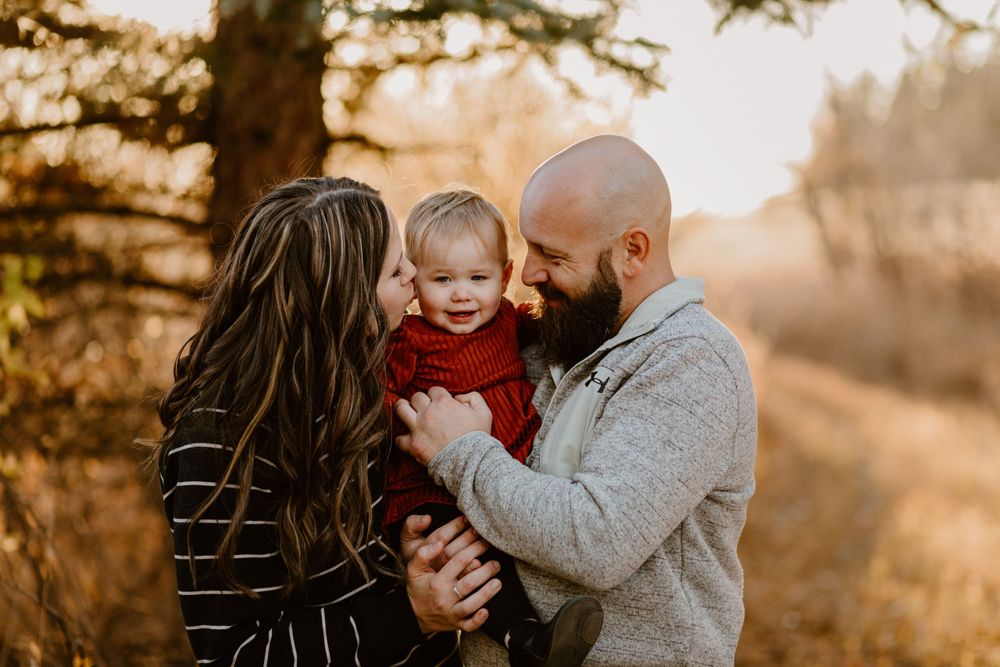 Golden sunset family portrait shows tender moment between parents and baby in red sweater outdoors.