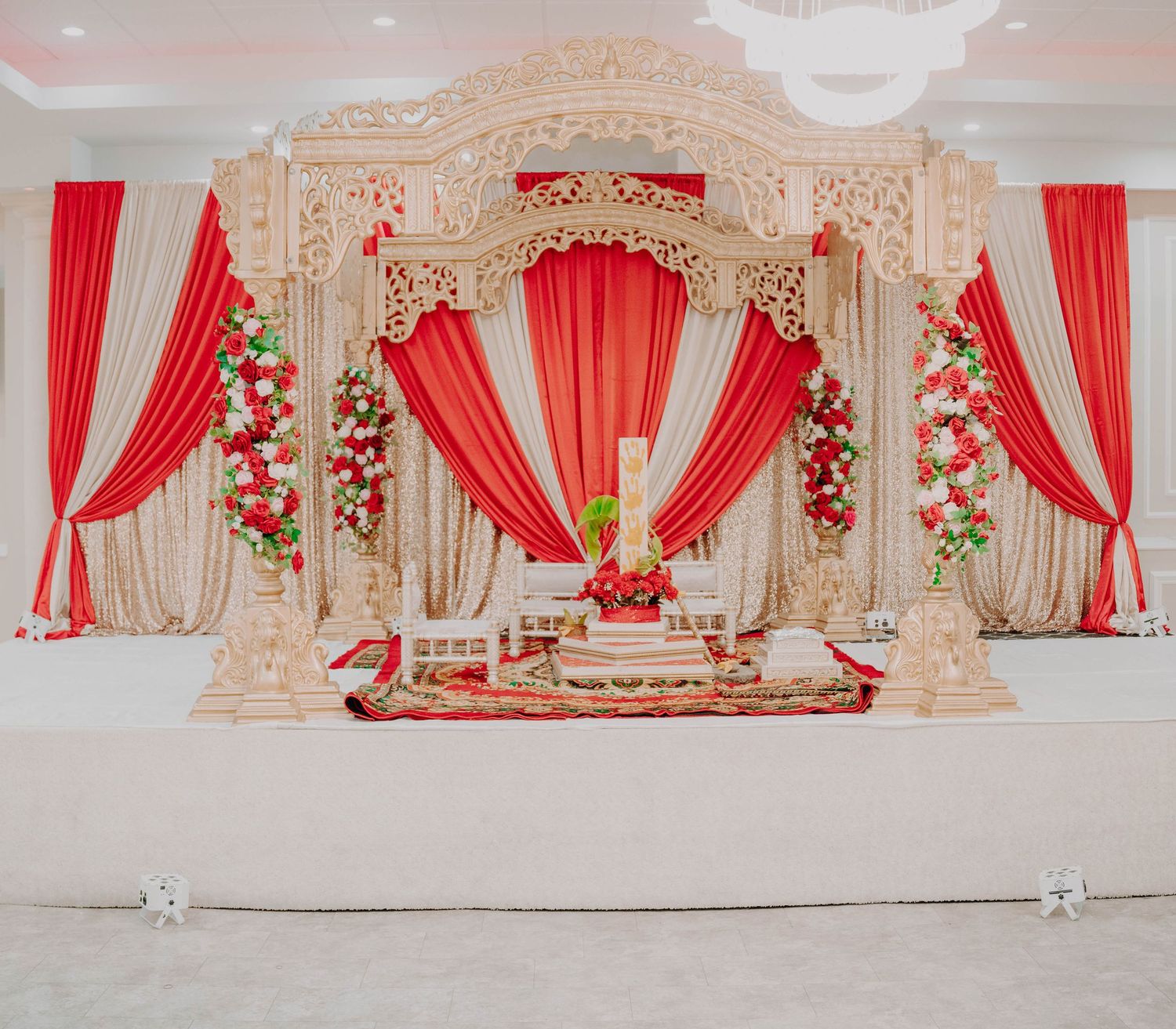 Elegant wedding mandap stage with ornate white columns and flowing red drapes decorated with floral arrangements.