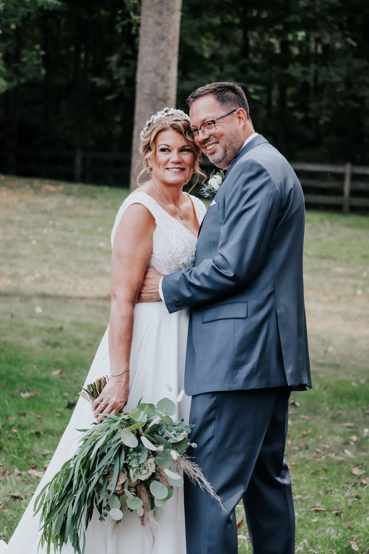 Wedding couple embraces outdoors while holding a lush greenery bouquet.