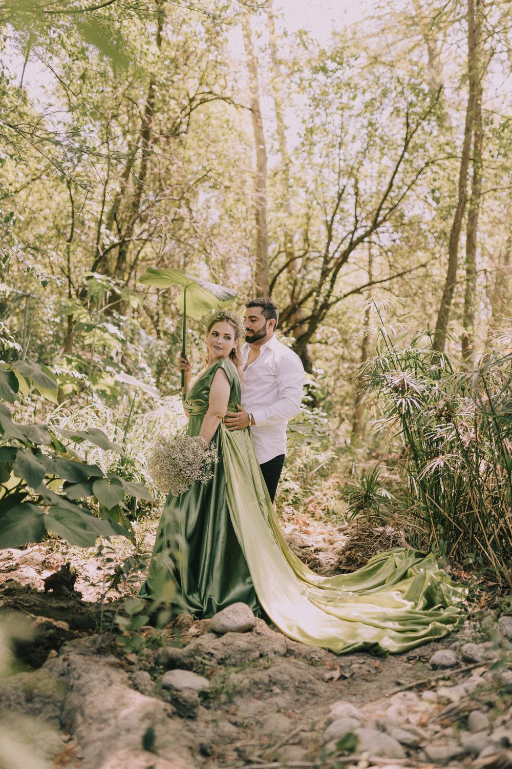 Couple in wedding attire poses dramatically among trees and fallen leaves.