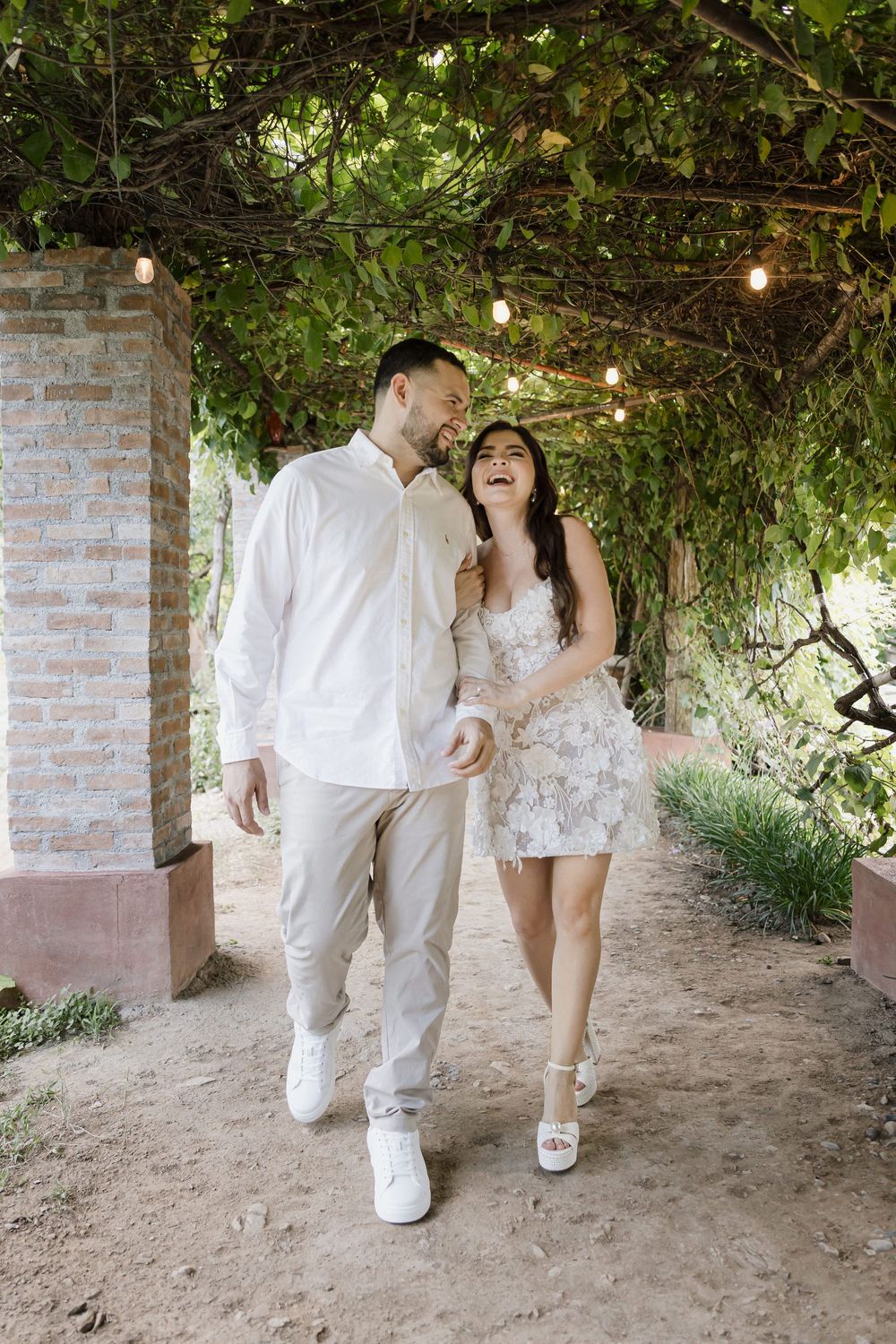 A couple shares a joyful moment under a vine-covered pergola with string lights.