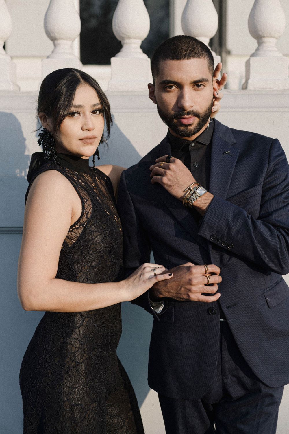 A stylish couple in black formal wear poses against white architectural columns.