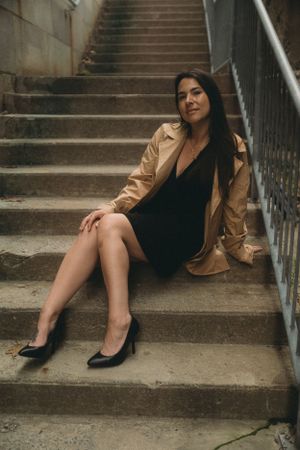 A woman in a trench coat and black dress poses elegantly on concrete steps.