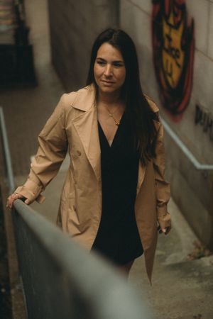 A woman in a beige trench coat stands confidently by a railing in an urban setting.