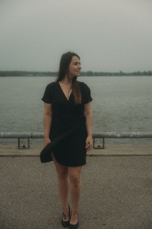 A woman in a black dress stands by a waterfront on an overcast day.