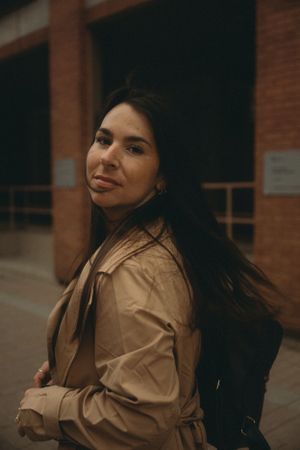 A woman in a beige trench coat poses against a brick building background.