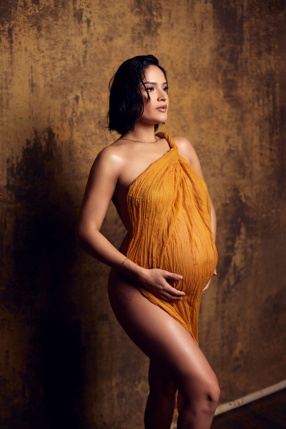Artistic maternity portrait in golden fabric against textured brown backdrop.