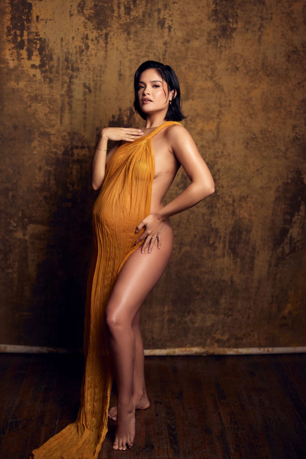 Dramatic maternity photography with flowing mustard fabric against rustic wall.