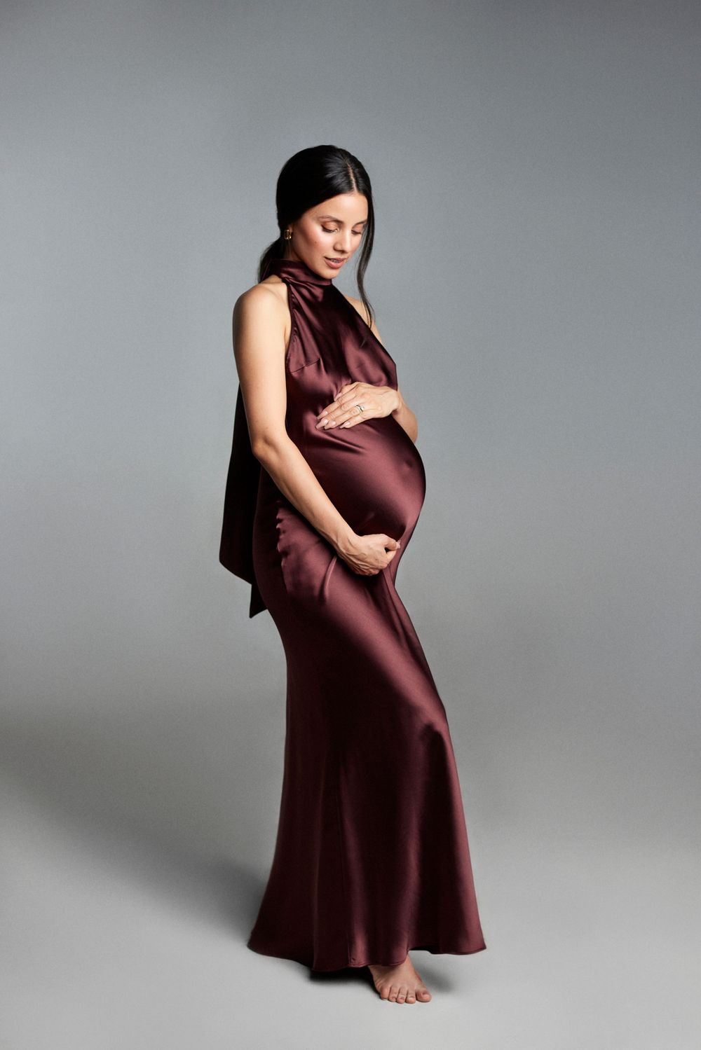 Maternity photography series featuring burgundy dress against grey studio background.