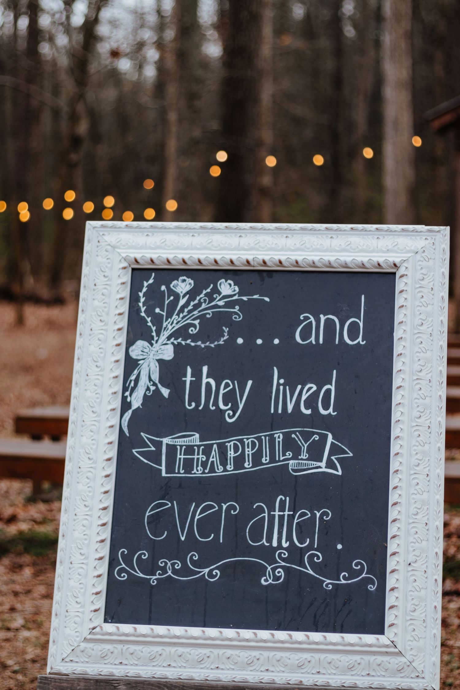 White-framed chalkboard sign with 'and they lived happily ever after' text against string lights.