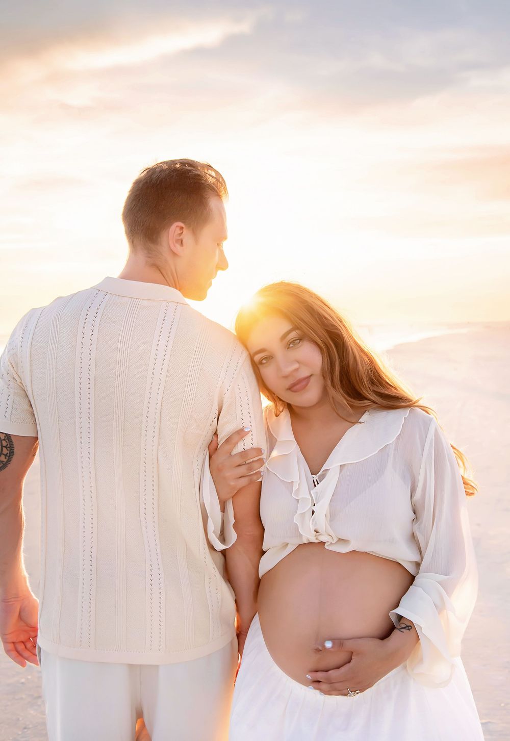 Pregnant couple embracing at sunset on white sand dunes, woman in white crop top cradling baby bump.